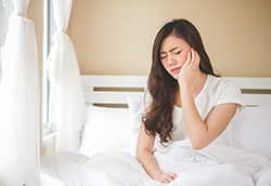 Woman sitting up in bed touching jaw in discomfort at Karl Hoffman Dentistry in Lacey, WA